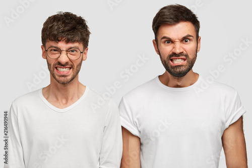 Studio shot of angry furious bearded men colleagues clench teeth in annoyance, feel irritation as recieve much work and duties from boss, dressed in casual white t shirts. Negative human emotions