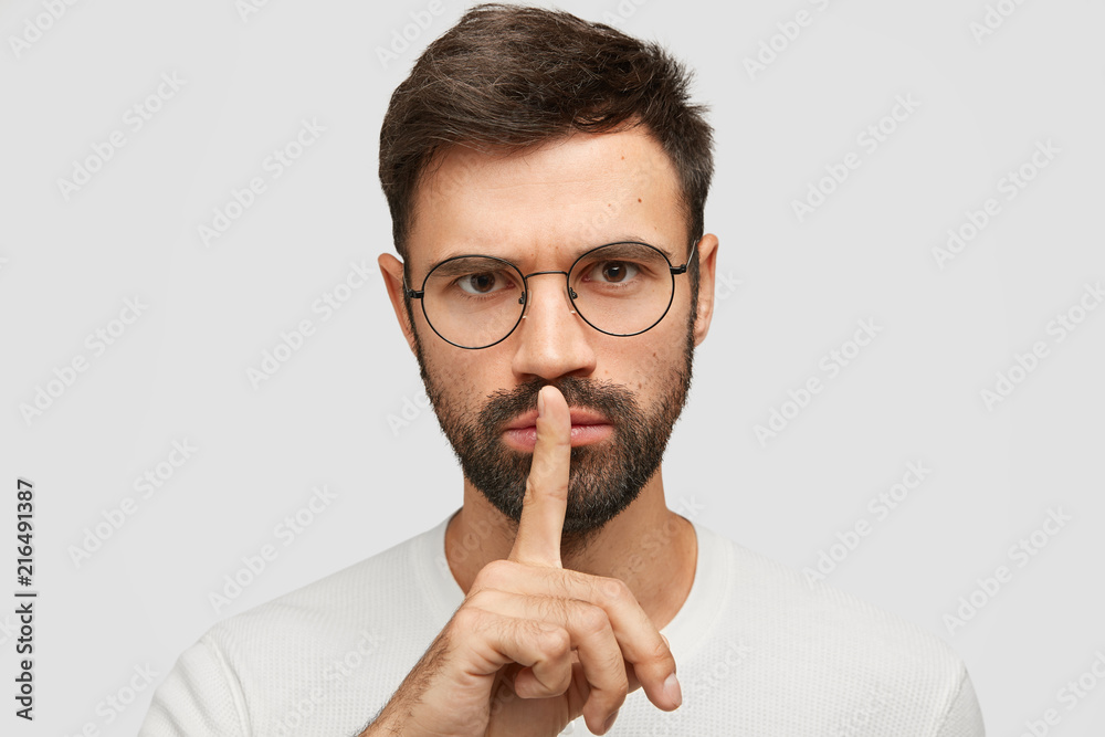 Headshot of handsome bearded Caucasian male makes silence gesture, looks seriously at camera, has concentrated look, demands complete silence, poses against white background. People and hush Headshot of handsome bearded Caucasian male makes silence gesture, looks seriously at camera, has concentrated look, demands complete silence, poses against white background. People and hush