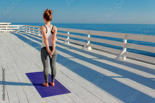 Young slim woman stretching outdoors at white wooden seafront. Girl standing on violet yoga mat with hands back in mudra. Exercise for shoulder blades