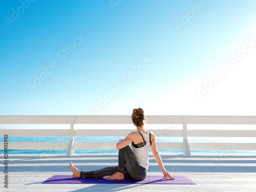 Young slim woman in tight sportswear practicing yoga outdoors at white wooden seafront. Sitting twisting pose. photo