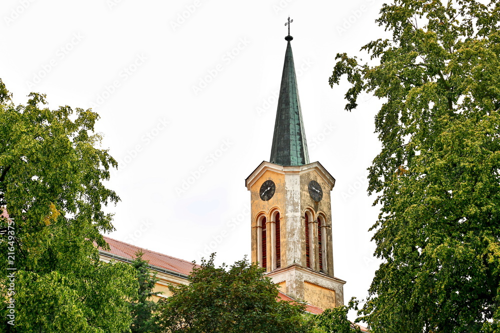 Fototapeta premium Church tower with green roof and a metal cross on the top, yellow and white paint-peeled facade, red roof, historical architecture, old clock, Saint Thomas in Hudlice, Czech Republic, Europe