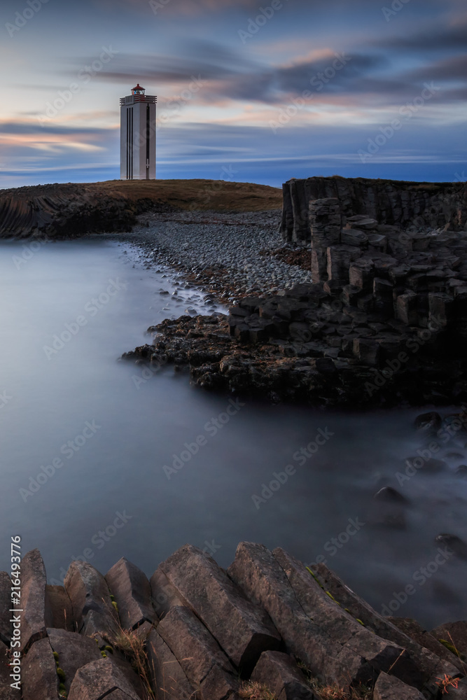 Fototapeta premium Leuchtturm im letzten Abendlicht. Im Vordergrund Basaltgestein. Im Norden Islands