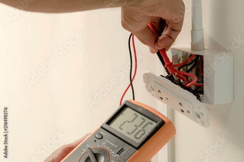 Electrician is using a digital meter to measure the voltage at the power outlet in on the wall.