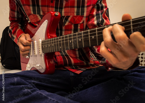 Young man sitting on the floor with his legs crossed playing a red electric guitar. Musician dressed in jeans and red plaid shirt. Using a pick.