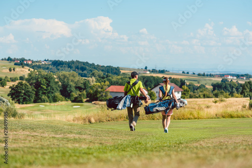 Full length rear view of a couple carrying professional golf bags, while walking on green field towards the golf course in a sunny day of summer