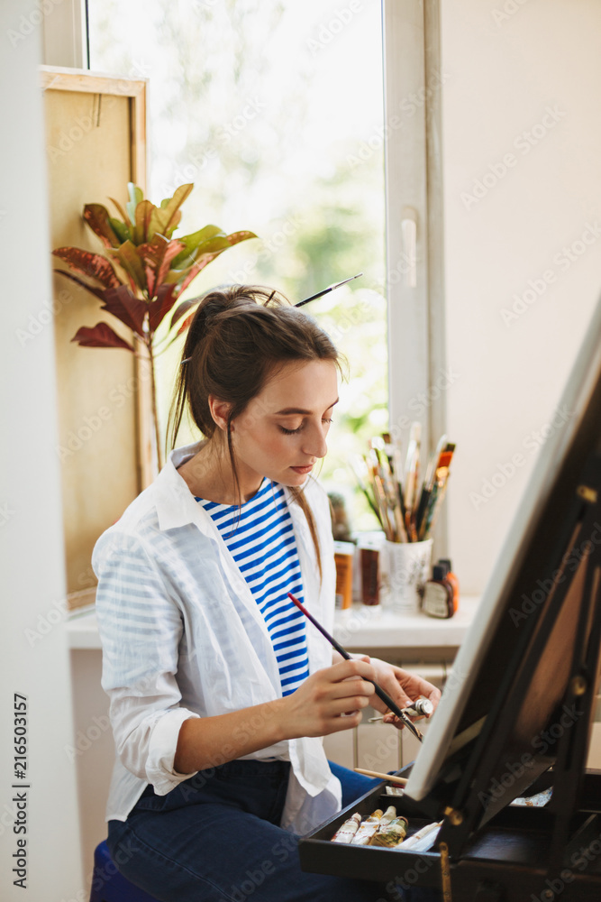 Beautiful girl in white shirt and striped T-shirt dreamily preparing to drawing on easel with window on background at home