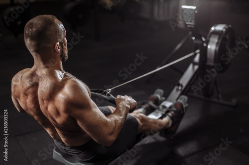 close up back view image. well-built guy is warming up with rowing machine. full length photo