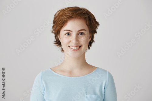 Portrait of attractive redhead caucasian female with freckles standing in blouse over gray background, smiling and gazing at camera with huge desire, hearing great offer and wanting to take it