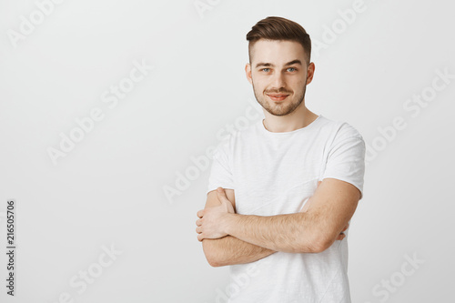 Intrigued smart young male entrepreneur in white t-shirt with moustache smirking holding hands crossed on chest feeling satisfied and glad receiving great result in sales over gray background