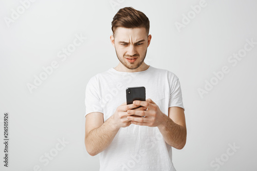 Wtf it mean. Perplexed questioned handsome guy with brown stylish hairstyle frowning confused while looking at smartphone screen reading strange disappointing message in device over gray wall © Cookie Studio