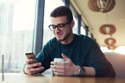 Young owner of coffee shop drinking cup of coffee looking forward writing SMS messages and communicate with friends in social networks via mobile phone using wifi internet connection and modern device photo