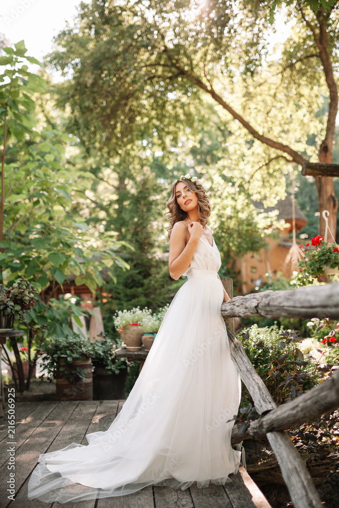 A cute curly woman in a white wedding dress with a wedding bouquet and wreath in her hair standing back to the camera in nature. Concept escaped bride. Forward to a happy bright future Runaway
