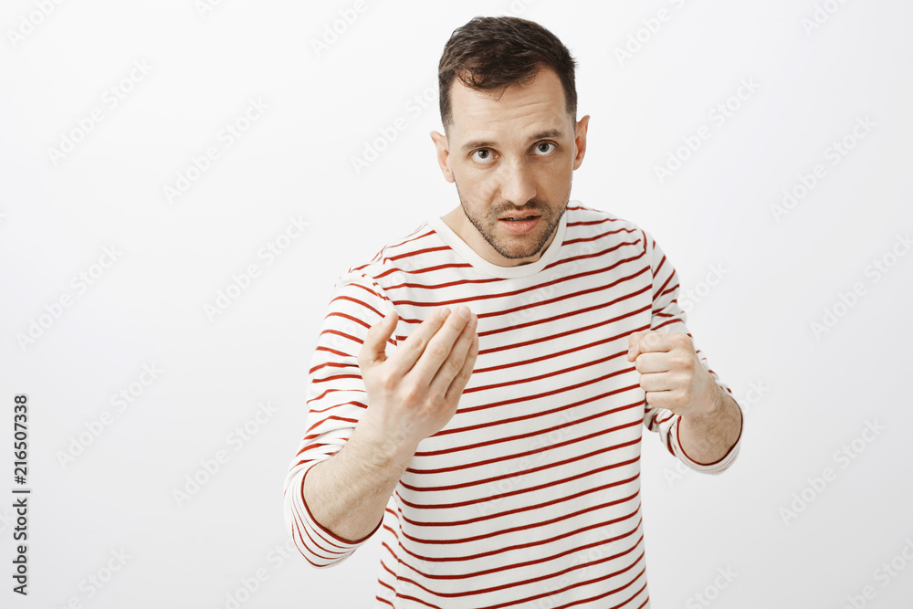 Fototapeta premium Your face pisses me off. Portrait of outraged angry adult male, showing come here gesture and raising fist, looking from under forehead with threatening look, wanting to fight or punch offender