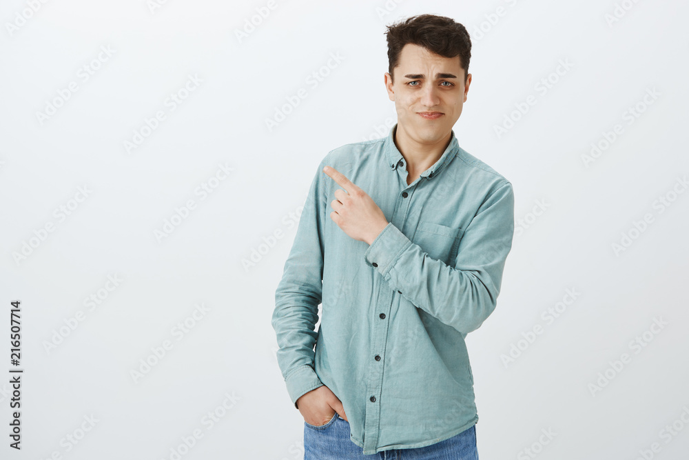 Indoor shot of unsure doubted good-looking european male, tilting right and pointing at upper left corner, frowning and grimacing from ugly disgusting thing, standing over gray background Indoor shot of unsure doubted good-looking european male, tilting right and pointing at upper left corner, frowning and grimacing from ugly disgusting thing, standing over gray background