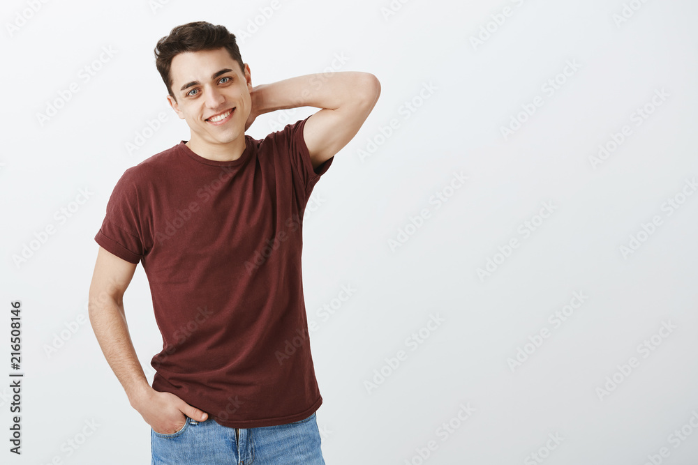 Shy good-looking masculine guy in red t-shirt, touching back of neck and holding hand in pocket, smiling broadly while receiving compliments or thanks, being timid and pleased over gray wall Shy good-looking masculine guy in red t-shirt, touching back of neck and holding hand in pocket, smiling broadly while receiving compliments or thanks, being timid and pleased over gray wall