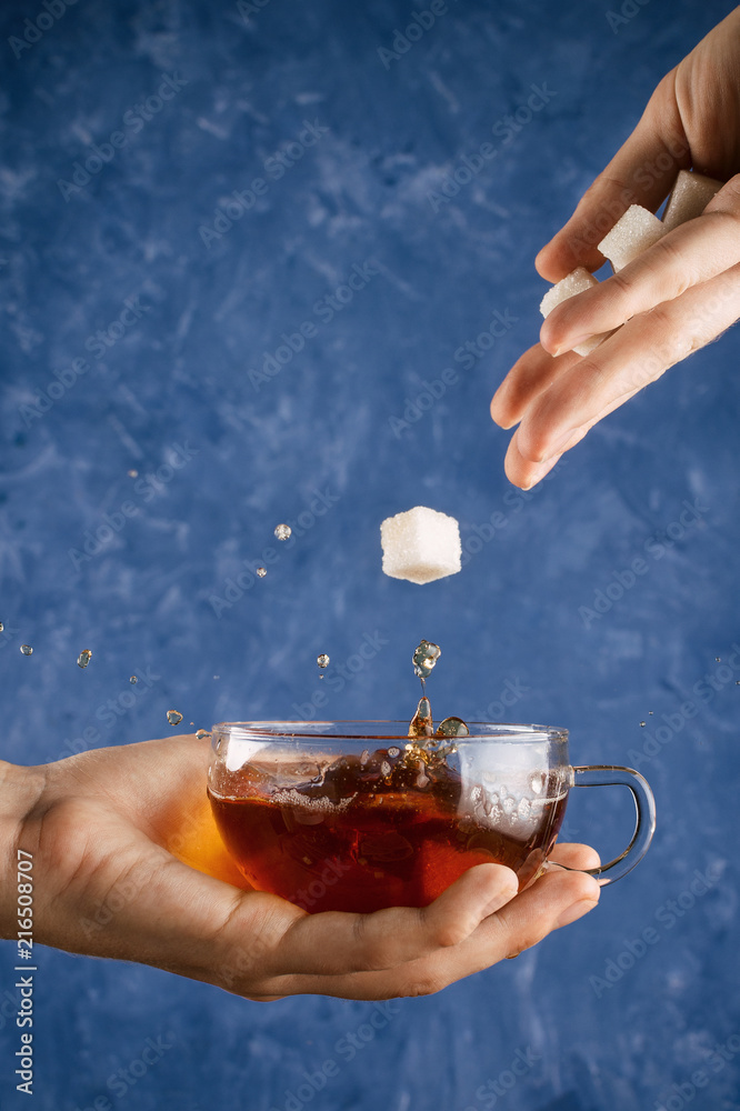 Woman's hands holding cup with warm splashing black tea and dropping sugar cubes into it, selective focus Woman's hands holding cup with warm splashing black tea and dropping sugar cubes into it, selective focus