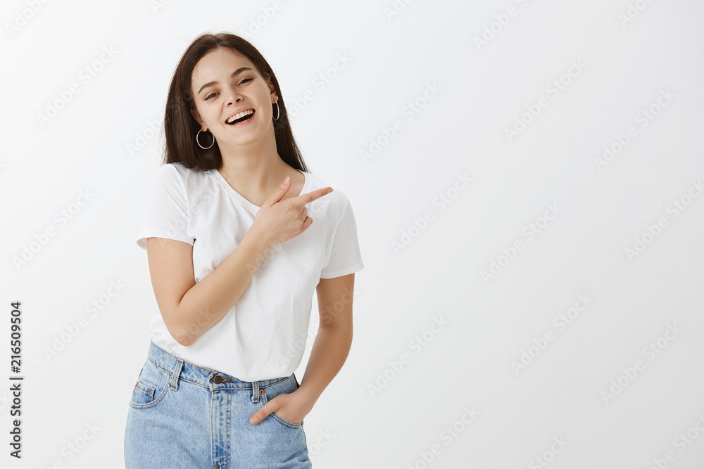 Charming hot and stylish dark-haired female in stylish street style outfit, holding hand in pocket and pointing right with forefinger, laughing joyfully, standing happily over gray background