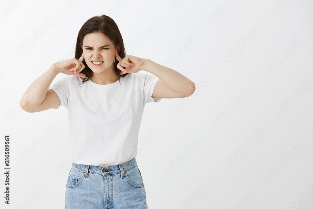 You play awful, I hate it. Irritated and annoyed attractive woman with dark hair, frowning and grimacing, covering ears with index fingers, hearing terrible noise, being pissed with loud sound