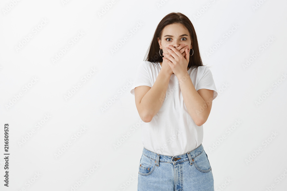Girl loosing speech being shocked to bottom of heart, gasping, covering mouth with palm, staring at camera stunned and amazed, standing against gray background under great impression over grey wall Girl loosing speech being shocked to bottom of heart, gasping, covering mouth with palm, staring at camera stunned and amazed, standing against gray background under great impression over grey wall