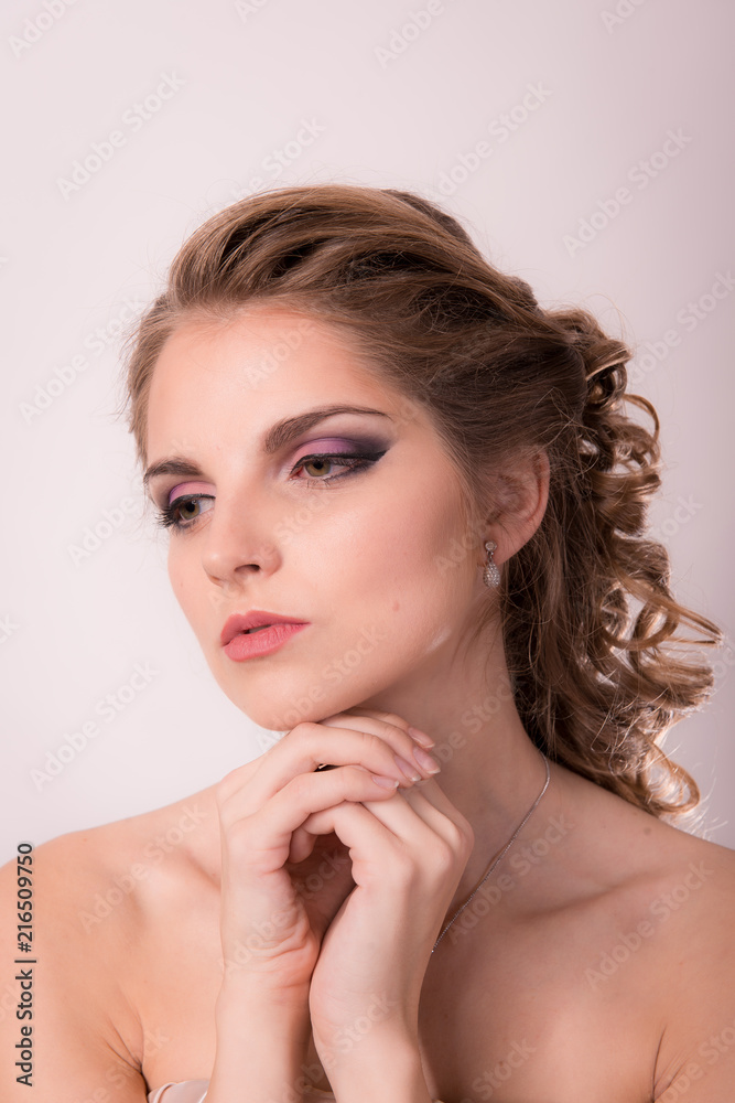 Brown-haired woman with a professional hairdress and make-up is holding hands near the face on a light background close-up.