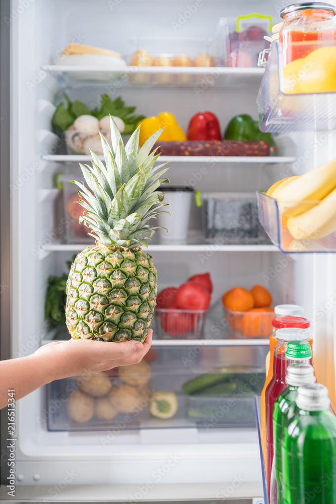 Hand with fresh pineapple against open fridge Hand with fresh pineapple against open fridge