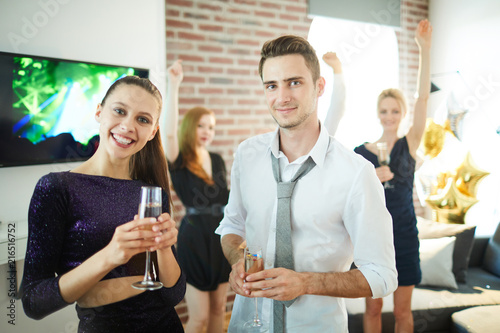 Happy young couple with champagne standig in front of camera on background of dancing friends
