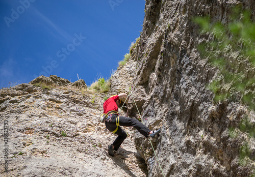 Photo from back of climbing young sports guy in yellow helmet on rock at summer day