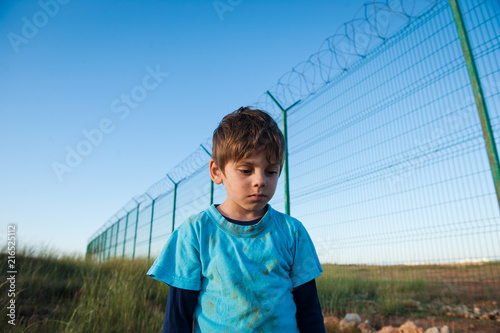 Obraz w ramie upset poor little boy refugee with dirty face near wall fence on border