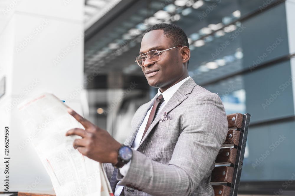 Fototapeta premium black man businessman in a business suit and glasses sits on a bench and reads a newspaper against the backdrop of a modern city