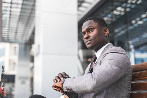 black man businessman in a business suit, expensive watch and glasses sitting on a bench and talking on the phone against the backdrop of a modern city