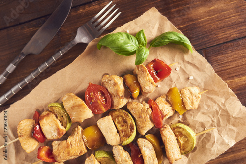 Bbq chicken breast with vegetables on wooden skewers on papper with spices and basil over wooden background.