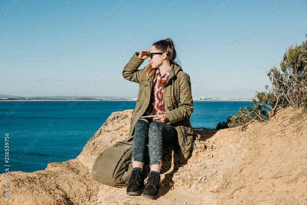 A young beautiful girl or tourist uses a tablet and looks into the distance. Ocean or sea in the background.