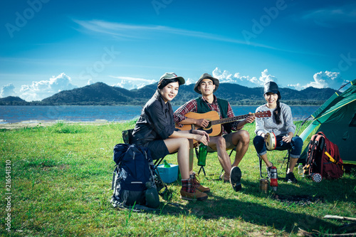 A  group of Asian friends sitting on chairs, singing, playing a guitar and drinking some beer and water together outside the tent near the fire while they has camping on Weekend holiday.