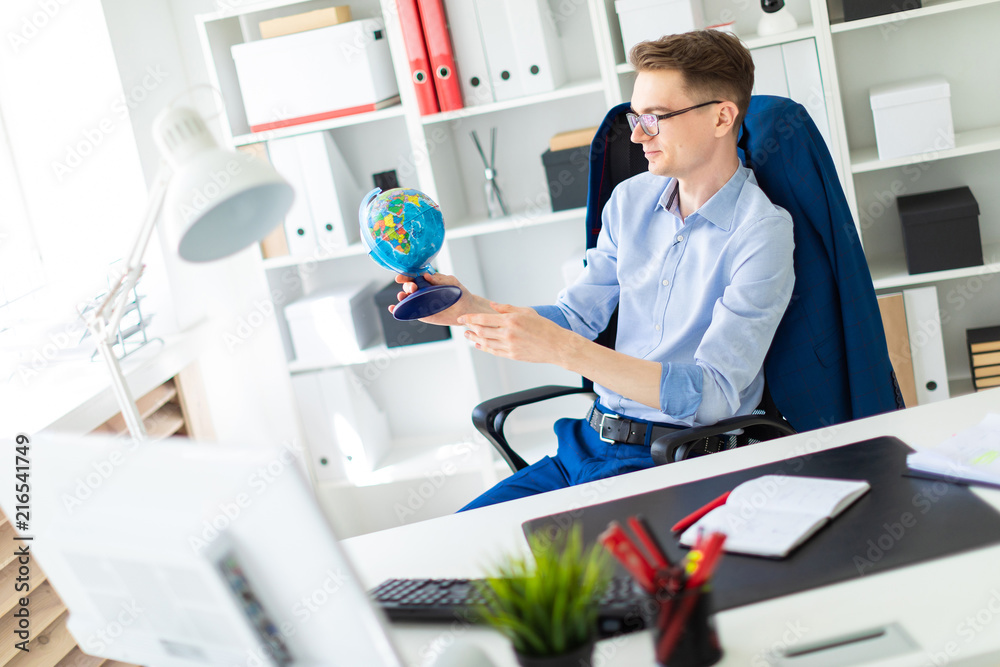 A young man sits in the office at a computer desk and holds a globe in his hands.