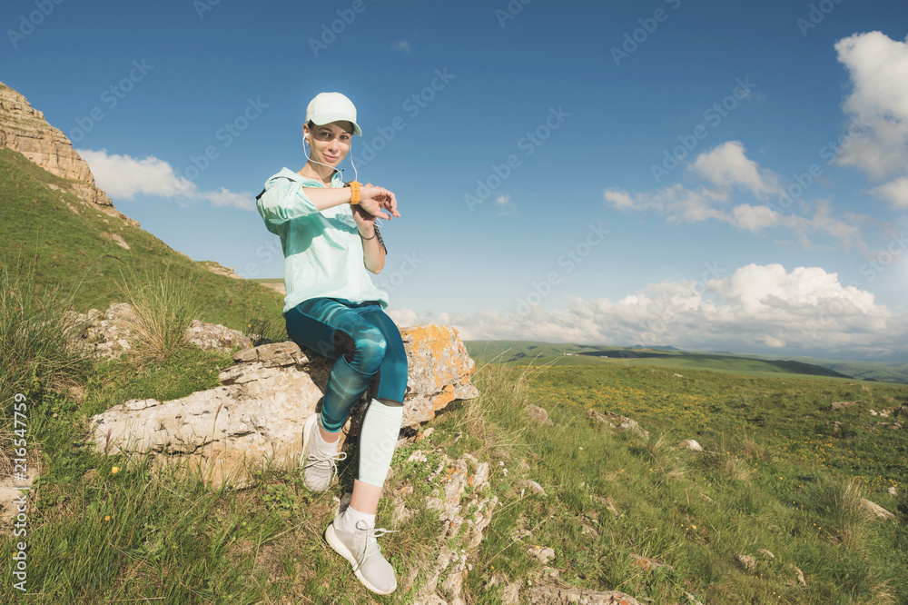 Portrait of a smiling young fitness girl in a cap and headphones checking her smart clock sitting on a rock outdoors against a blue sky with clouds Portrait of a smiling young fitness girl in a cap and headphones checking her smart clock sitting on a rock outdoors against a blue sky with clouds