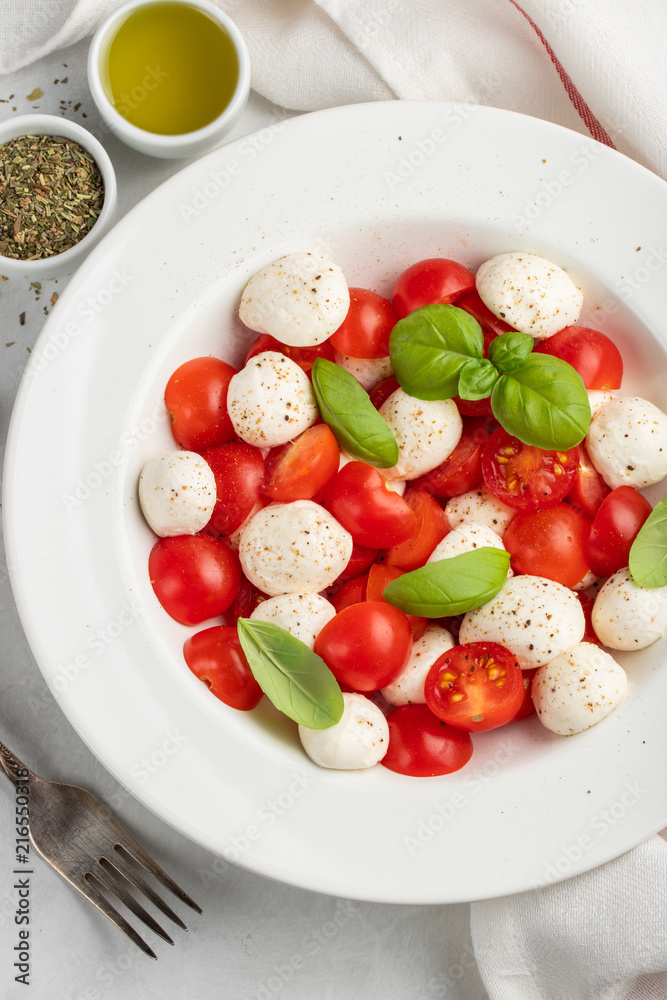 White plate of classic delicious caprese salad with ripe tomatoes, mozzarella and fresh basil leaves on gray concrete background. Italian food. The concept of a healthy vegetarian diet. Top view White plate of classic delicious caprese salad with ripe tomatoes, mozzarella and fresh basil leaves on gray concrete background. Italian food. The concept of a healthy vegetarian diet. Top view