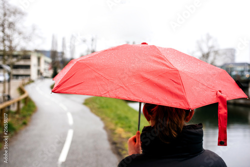Fototapeta Naklejka Na Ścianę i Meble -  Rear view of woman with red umbrella walking in city on a warm spring day - book cover and cinematic