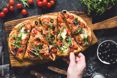 Flatbread pizza garnished with fresh arugula on wooden pizza board, top view. Dark stone background. Person picking slice of pizza
