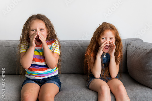 Little girls sitting on sofa screaming with hands around mouth photo