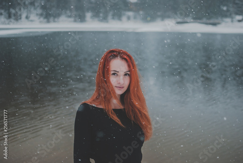 Joyful handsome black-eyed redhead girl looking at the camera. Splendid shady black mountain lake reflecting trees of forest behind her back.