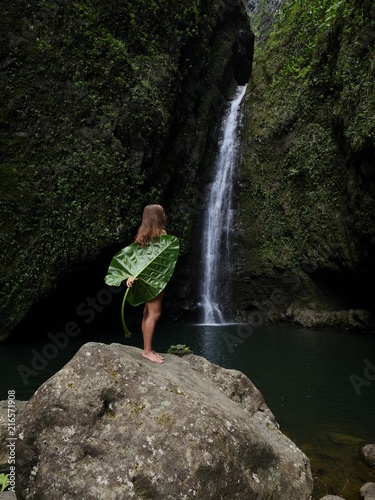 woman covered with big green leaf standing on a rock at a tropical Hawaiian waterfall sacred falls Oahu island 