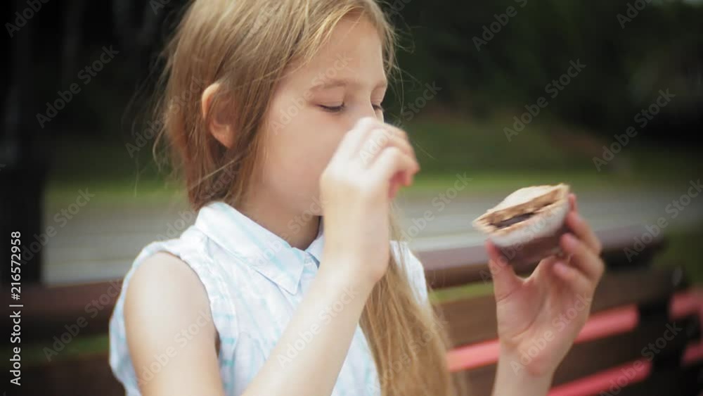 Close up of adorable little girl eating a cake with her hands sitting on a bench in the park Close up of adorable little girl eating a cake with her hands sitting on a bench in the park