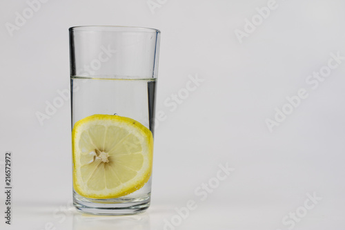 A glass of cold water with lemon on a white kitchen table. A refreshing drink in warm weather.