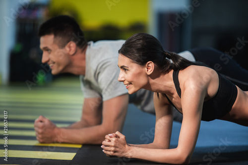 Focus on cheerful woman strained while doing plank. She is exercising with male friend and they are doing same static exercise while balancing on elbows. Two athletes are enjoying joint workout and