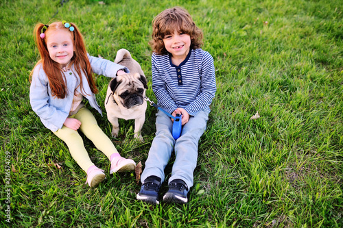 baby boy with curly hair and girl with red hair sitting on grass in park with dog pug photo