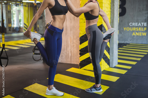 Shapely female couple are training flexibility after suspension workout. They are standing face to face and helping each other by keeping friend shoulder with hand. Women are standing on one foot and