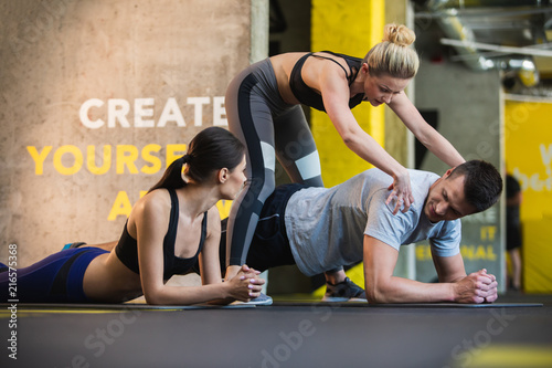 Woman instructor is assisting two people during workout in gym. She is explaining correct fulfilment of plank for guy while standing over him. His girlfriend is watching them and listening to
