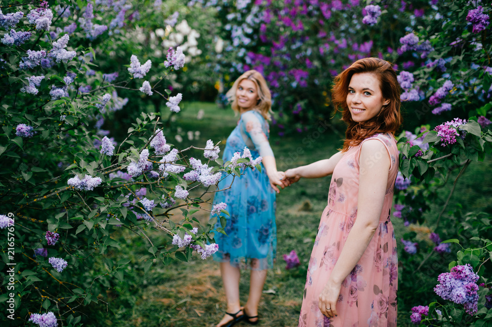 Romantic mood. Twin sisters tender relationship. beautiful twins young models in colorful summer dresses posing for camera in blooming lilac garden. Cheerful cute stylish girls in magic park outdoor. Romantic mood. Twin sisters tender relationship. beautiful twins young models in colorful summer dresses posing for camera in blooming lilac garden. Cheerful cute stylish girls in magic park outdoor.