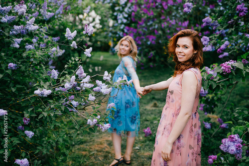 Romantic mood. Twin sisters tender relationship. beautiful twins young models in colorful summer dresses posing for camera in blooming lilac garden. Cheerful cute stylish girls in magic park outdoor.