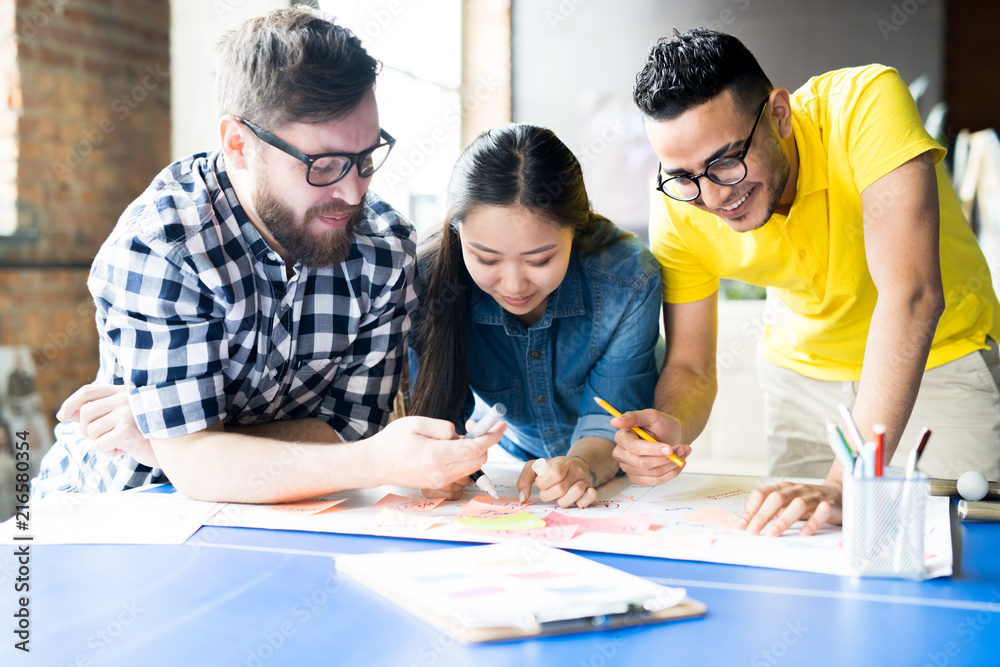 Naklejka premium Portrait of three creative young business people making up plan chart while standing at desk in modern office
